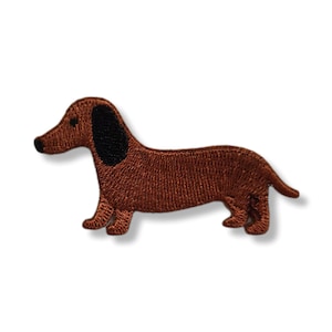 Może przedstawiać: Haftowana naszywka przedstawiająca brązowego jamnika z czarnymi uszami. Naszywka przedstawia psa z profilu, podkreślając jego długie ciało i krótkie nogi. Odpowiednia do odzieży lub akcesoriów.