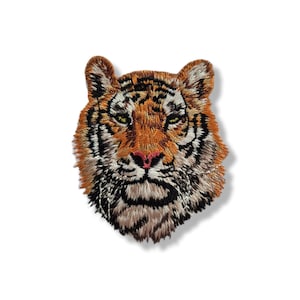 Puede incluir: Parche bordado con la cabeza de un tigre. El parche muestra costuras detalladas en tonos naranja, negro y blanco, con una nariz rosa y ojos amarillos. La cara del tigre está centrada sobre un fondo blanco.