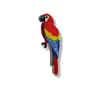 May include: Embroidered patch of a vibrant parrot. The bird features a red head and body, with yellow, blue, and green accents on its wings and tail. The parrot is perched on a black branch, with a white background.