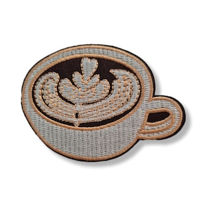 May include: An embroidered patch shaped like a coffee cup viewed from above. The cup contains dark brown coffee with light grey and tan latte art in a leaf pattern. The cup itself is light grey with a tan rim and a small handle on the right side.