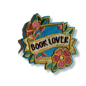 May include: Embroidered patch with a teal background and a banner that reads "BOOK LOVER." The patch features pink flowers, green leaves, and gold accents. The design is reminiscent of a traditional tattoo.