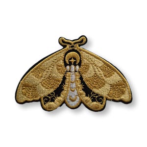 May include: Embroidered moth patch in gold, black, and white. The moth features detailed wing patterns and a crescent moon and cross design on its head. The patch is likely for clothing or accessories.