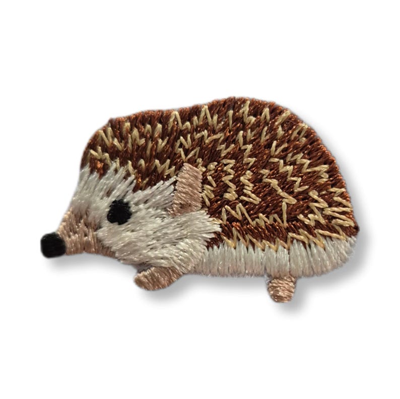 May include: Embroidered hedgehog patch in shades of brown, beige, and cream. The patch has a detailed design with a textured appearance, showcasing the hedgehog's quills and body. The hedgehog has black eyes and a small nose.