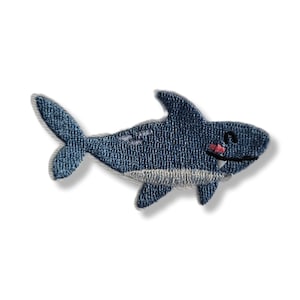 Peut inclure: Écusson brodé représentant un requin souriant en denim bleu. Le requin a un ventre blanc, une bouche rose et des yeux noirs. Idéal pour ajouter une touche marine et amusante aux vêtements ou accessoires.