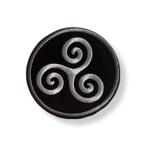 Op de afbeelding: Een geborduurde zwarte en witte patch met een rond ontwerp met een zilverkleurig driedubbel spiraal symbool.