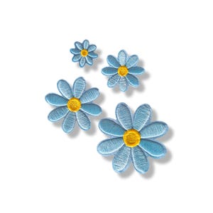 Puede incluir: Cuatro parches bordados en forma de flor de diferentes tamaños, cada uno con pétalos de color azul claro y un centro circular de color amarillo brillante. Los parches están dispuestos en grupo, mostrando una textura cosida detallada en los pétalos.