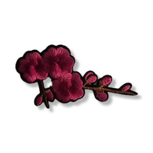 Puede incluir: Parche bordado con tres flores de color rosa intenso con borde negro, unidas a un tallo marrón con capullos. El parche está diseñado para planchar o coser sobre tela, añadiendo un elemento floral decorativo.