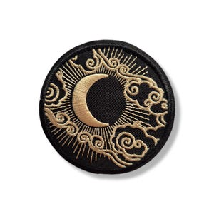 Op de afbeelding: Ronde zwarte patch met een gouden halve maan ontwerp. De maan is omgeven door gouden stralen en wervelende wolkenachtige patronen. De patch is geborduurd met een gedetailleerd, hemels thema.