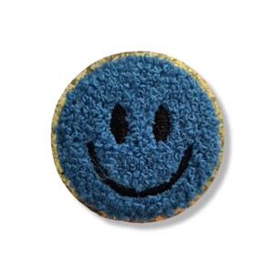 May include: A circular coaster featuring a textured blue tufted surface with a black embroidered smiley face design. The base of the coaster is made of natural light brown cork, visible around the edges of the blue tufted circular top.
