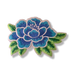 Könnte beinhalten: Gestickter Blumen-Patch mit einer blauen Blume in verschiedenen Blau- und Türkistönen, umgeben von grünen Blättern. Die Blume hat einen hellrosa Rand, ideal für Kleidung oder Accessoires.