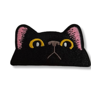 May include: A black cat face patch with yellow eyes, pink ears, and a brown nose. The patch is embroidered with a detailed design, perfect for adding to clothing or accessories. The cat's face is a simple, yet charming design.