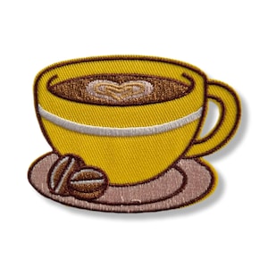May include: Embroidered patch of a yellow coffee cup with a white stripe and a brown handle. The cup contains coffee with a heart design. Two coffee beans sit on the saucer. The patch is on a white background.