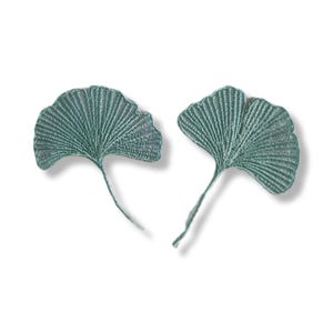 Könnte beinhalten: Zwei teal-farbene, gestickte Ginkgoblatt-Applikationen. Jedes Blatt hat eine fächerartige Form mit detaillierten Adern und einem schlanken Stiel. Die Blätter sind vor einem weißen Hintergrund platziert.