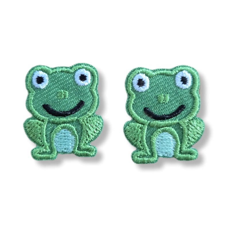 May include: Two identical embroidered patches featuring a cartoon frog design. Each patch displays a bright green frog with large white eyes, black pupils, a curved black mouth, and a light blue belly, all outlined with detailed green stitching.