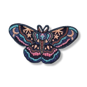 May include: An embroidered patch shaped like a moth with a dark navy blue base. The design features intricate stitching in shades of light pink, turquoise, and peach, depicting celestial motifs including crescent moons and stars across the wings.