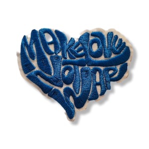 Può includere: Toppa ricamata a forma di cuore con le parole "Make Love Happen" in filo blu. La toppa ha uno sfondo color crema e un bordo bianco. Il testo è in un carattere corsivo stilizzato.