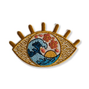 Peut inclure: Patch brodée thermocollante représentant un œil stylisé avec une vague, un soleil et des fleurs de cerisier à l'intérieur. L'œil est bordé d'or sur un fond blanc.