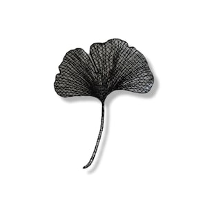 Può includere: Un'opera d'arte da parete a forma di foglia di ginkgo in metallo nero. La foglia è finemente progettata con un motivo a rete, e il gambo è una linea sottile e strutturata. L'opera è ambientata su uno sfondo bianco.