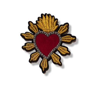 Op de afbeelding: Geborduurde patch met een rood hart, zilveren rand, gouden stralen en een zwarte omtrek. Het gedetailleerde ontwerp is geschikt voor kleding of accessoires. Het is een decoratief item.