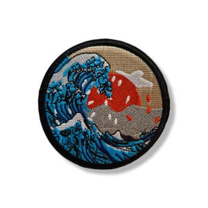 Peut inclure: Écusson rond brodé représentant une vague japonaise stylisée avec un soleil rouge, une montagne grise et des vagues bleues. Le patch a une bordure noire et un fond beige.