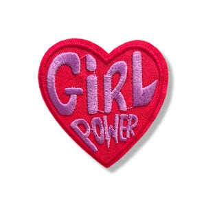 Op de afbeelding: Een hartvormige geborduurde patch met een felrode achtergrond en een gestikte rand. De tekst "GIRL POWER" is in lichtpaars garen in het midden van het hart geborduurd, met een gedurfd, gestileerd lettertype met een getextureerde afwerking.