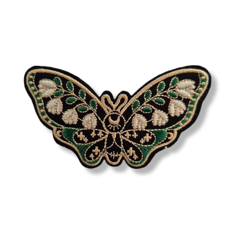 Peut inclure: Un écusson brodé en forme de papillon sur une base en tissu noir. Le motif présente des fleurs de muguet couleur crème, des feuilles vertes et des détails en fil doré, incluant un symbole de croissant de lune et d'œil au centre.