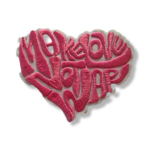 May include: A pink embroidered heart-shaped patch with the words "Make Love". The patch is stitched onto a light beige fabric. The cursive font is a prominent feature.