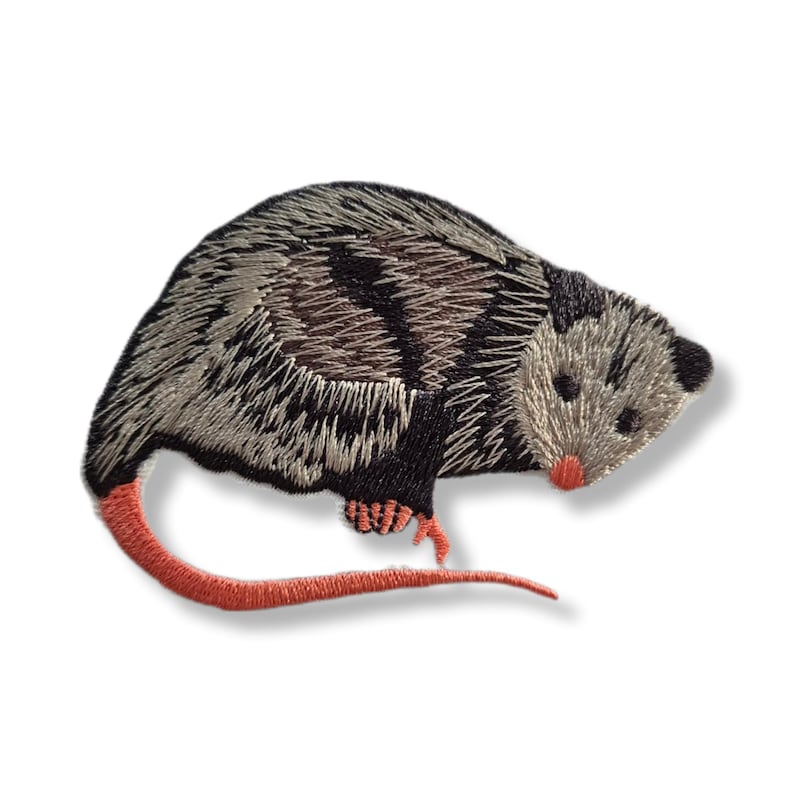May include: Embroidered opossum patch. The patch depicts an opossum with grey and black fur, a pink nose, feet, and tail. This patch is designed to be ironed or sewn onto fabric, ideal for customising clothing or accessories.
