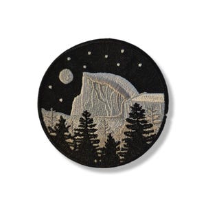 Op de afbeelding: Ronde geborduurde patch met een nachtelijk berglandschap. Het ontwerp toont een lichte berg, donkere bomen, een maan en sterren tegen een zwarte achtergrond. Geschikt voor kleding of tassen.