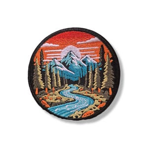 Puede incluir: Un parche bordado circular que presenta una escena de paisaje con un río azul serpenteante en primer plano, pinos de color verde oscuro y montañas azules cubiertas de nieve bajo un cielo naranja y rojo al atardecer.