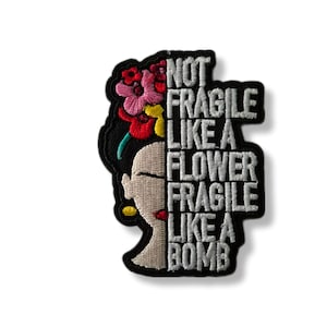 Peut inclure: Patch brodé avec une image stylisée de Frida Kahlo avec des fleurs dans les cheveux. Le texte "Not Fragile Like A Flower Fragile Like A Bomb" est écrit en blanc sur fond noir.