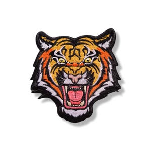 Op de afbeelding: Een geborduurde patch met de kop van een tijger die brult met open bek. Het ontwerp gebruikt levendige oranje, gele en witte draden met zwarte omlijningen en details, met scherpe tanden, een roze tong en doordringende gele ogen.