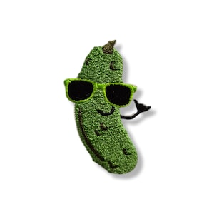 May include: Embroidered patch featuring a smiling green pickle wearing black sunglasses. The pickle has a small arm raised in a friendly gesture. The patch is on a white background.