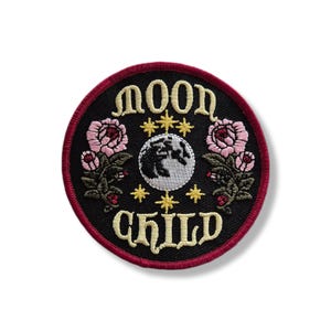 Könnte beinhalten: Ein runder, schwarzer Aufnäher mit den Worten "MOON CHILD" in goldener Schrift. Das Design zeigt eine Erdkugel, goldene Sterne und rosa Rosen. Der Aufnäher hat einen roten Rand.