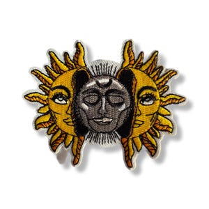 Peut inclure: Écusson brodé avec un motif céleste. Le design présente un visage de lune gris central flanqué de deux visages de soleil jaunes. Les soleils ont des traits détaillés et des rayons rayonnants. L'écusson est sur fond blanc.