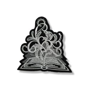 Può includere: Toppa ricamata in bianco e nero raffigurante un libro aperto con motivi floreali a vortice. La toppa ha uno sfondo nero e un ricamo bianco dettagliato. Il design ricorda il pizzo vintage.