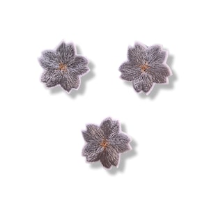 Può includere: Tre toppe ricamate a forma di fiori di ciliegio. Ogni toppa presenta un filo grigio argento con un bordo viola chiaro e un piccolo centro marrone dorato. I petali hanno un distinto design a cinque lobi con una trama di cucitura visibile.