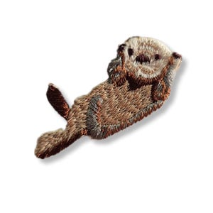 Op de afbeelding: Gestikt pleister van een bruine en witte zeeotter met een gekrulde staart, drijvend op zijn rug.