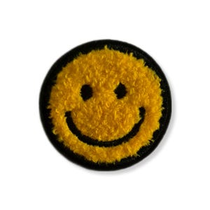 Puede incluir: Un parche redondo amarillo con la cara de un smiley con una textura difusa. El smiley tiene ojos negros y una boca negra curvada, con un borde negro. Este parche se puede coser o planchar.