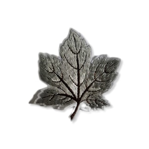 Puede incluir: Una hoja de arce bordada en plata con vetas marrones. La hoja es un parche o adorno decorativo.