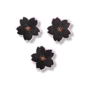 Könnte beinhalten: Drei gestickte Blumenaufnäher in Form von Kirschblüten. Jeder Aufnäher hat schwarze Blütenblätter mit einem hellrosa Rand und ein zentrales Bündel kleiner, beigefarbener Staubgefäße. Die Stickerei erzeugt eine strukturierte Optik.