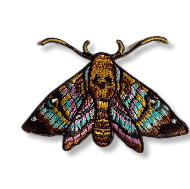 K&ouml;nnte beinhalten: Ein bestickter Aufn&auml;her in Form eines Nachtfalters mit einem Totenkopfmuster auf dem Thorax. Die Fl&uuml;gel zeigen eine lebendige Farbpalette aus t&uuml;rkisfarbenen, violetten und goldenen F&auml;den auf dunklem Grund, mit zwei langen Antennen am Kopf.