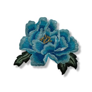 Peut inclure: Écusson brodé représentant une grande fleur bleue avec de multiples couches de pétales et un centre doré. La fleur est accompagnée de deux feuilles vertes. Le patch est conçu pour être cousu ou repassé sur du tissu.