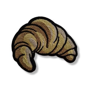 Op de afbeelding: Geborduurde patch van een croissant, met tinten bruin en geel. De patch heeft een zwarte rand en het ontwerp is gedetailleerd met getextureerde stiksels. Geschikt voor kleding, tassen of andere stoffen items.