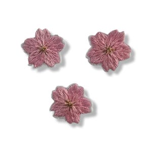 以下が含まれることがあります： 五枚花びらのデザインの、刺繍されたピンク色の花のパッチが3つ。 花は金色の中心部を持ち、白い背景に対して、テクスチャ加工された外観をしています。 これらのパッチは、生地の装飾に最適です。