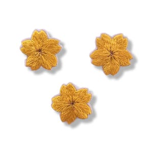 May include: Three individual embroidered patches shaped like five-petaled flowers. Each patch features a vibrant golden yellow thread color with a dense, textured stitch pattern radiating from the center of the floral design to the edges.