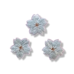 Peut inclure: Trois écussons brodés en forme de fleur de couleur bleu clair avec cinq pétales chacun. Le centre de chaque fleur présente un petit détail cousu marron. Les bords des pétales sont soulignés par une fine bordure rose clair sur fond blanc.