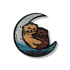 May include: Embroidered patch featuring two otters cuddling on a crescent moon. The otters are brown and tan, with the moon in silver and the water in shades of blue. The patch is designed for clothing or accessories.