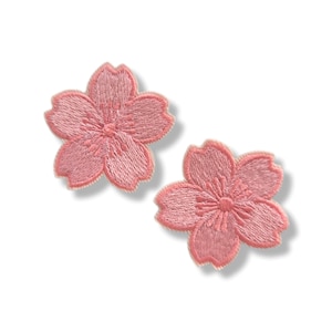以下が含まれることがあります： 5枚の花びらを持つ桜の花の形をした刺繍ワッペン2枚。各ワッペンは淡いピンク色の糸で構成され、花びらと中央の雄しべには濃いピンク色のステッチが施されており、白い背景の上に立体的な花の模様が表現されています。