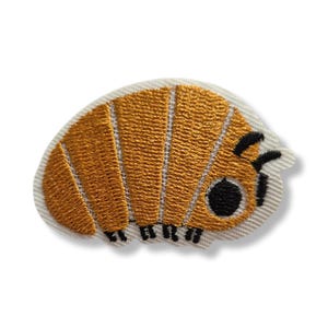 Pode incluir: Um emblema bordado em forma de tatu-bola, apresentando um corpo segmentado amarelo-dourado com contornos brancos, um grande olho circular preto e pequenas antenas e patas pretas, sobre um fundo de tecido branco com borda recortada.